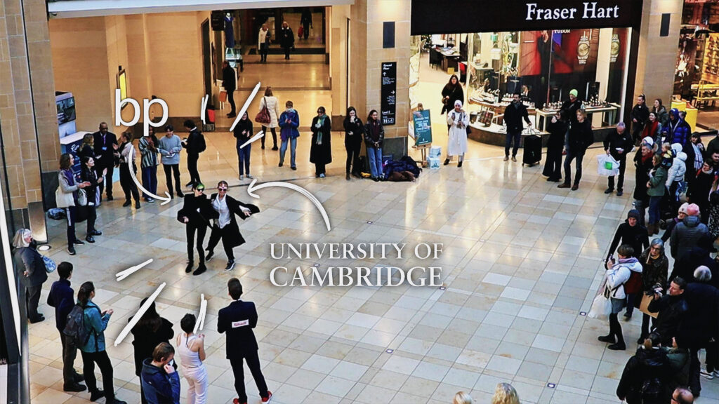 Composite still from flashmob in Grand Arcade showing 2 CCJ members, with the logos of bp and the University of Cambridge on their forehead respectively. They are dancing together with their arms linked to symbolise the close collaboration between the 2 institutions. 